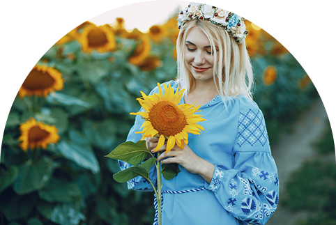 Woman in a blue embroidered dress holding a sunflower and smiling in a blooming sunflower field, wearing a floral crown.