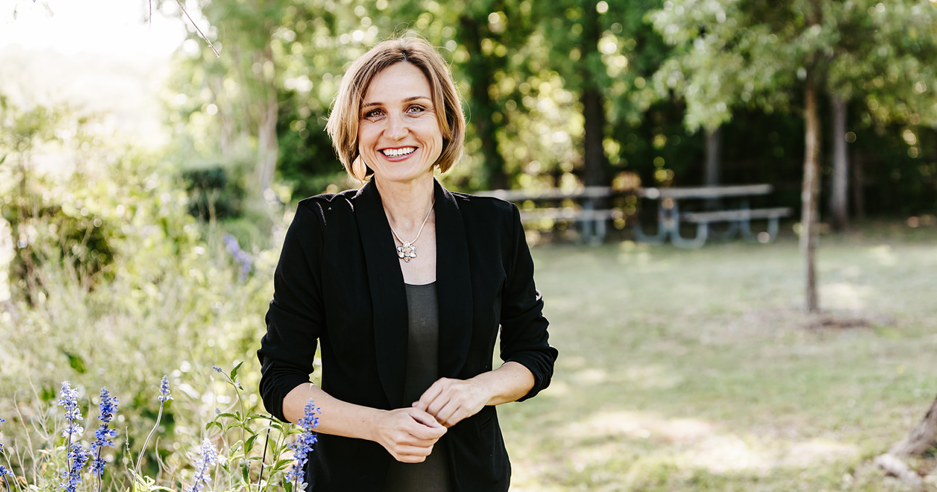 Lana Savit standing confidently in a garden, smiling with arms crossed, wearing a black blazer, symbolizing empowerment, professionalism, and holistic leadership for women over 40.