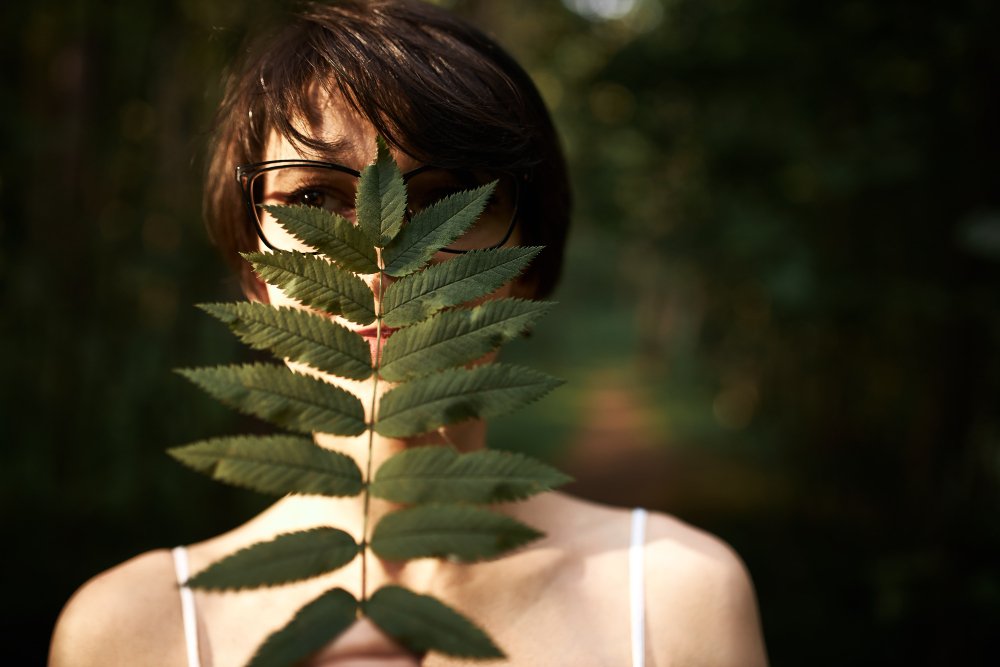 Woman partially hidden behind a fern leaf in a forest, symbolizing natural connection, introspection, and holistic self-discovery.