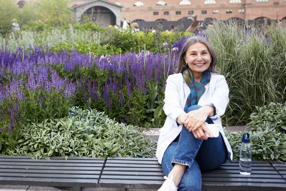 Smiling older woman sitting on a bench in a lush garden with purple flowers, symbolizing vibrant aging, wellness, and connection to nature.